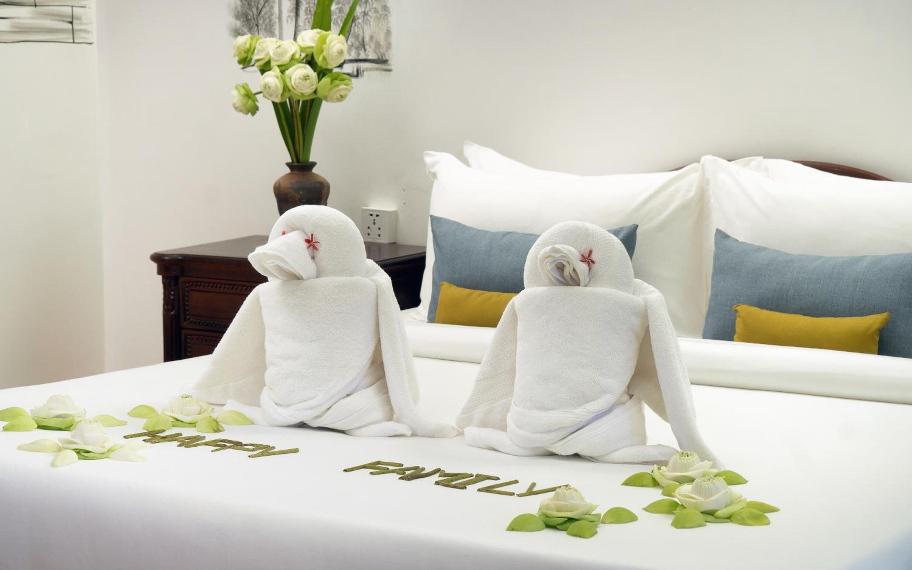 A hotel room with a bed, two towels with red flowers, a vase with white flowers, and a 'Happy Family' decoration on the bed.