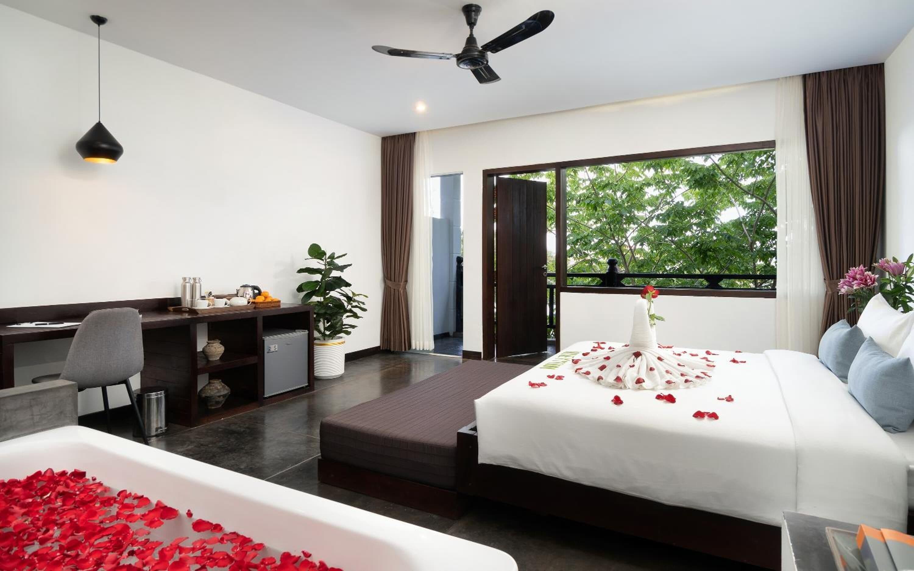 A hotel room with a bed, a desk, a chair, a ceiling fan, and a window with a view of trees.