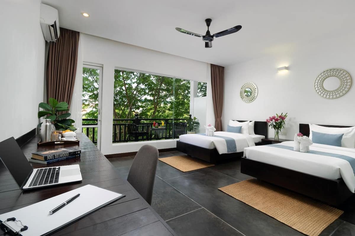 Habitación de hotel moderna con dos camas, un escritorio con una computadora portátil, un ventilador de techo y un balcón con vistas a árboles.