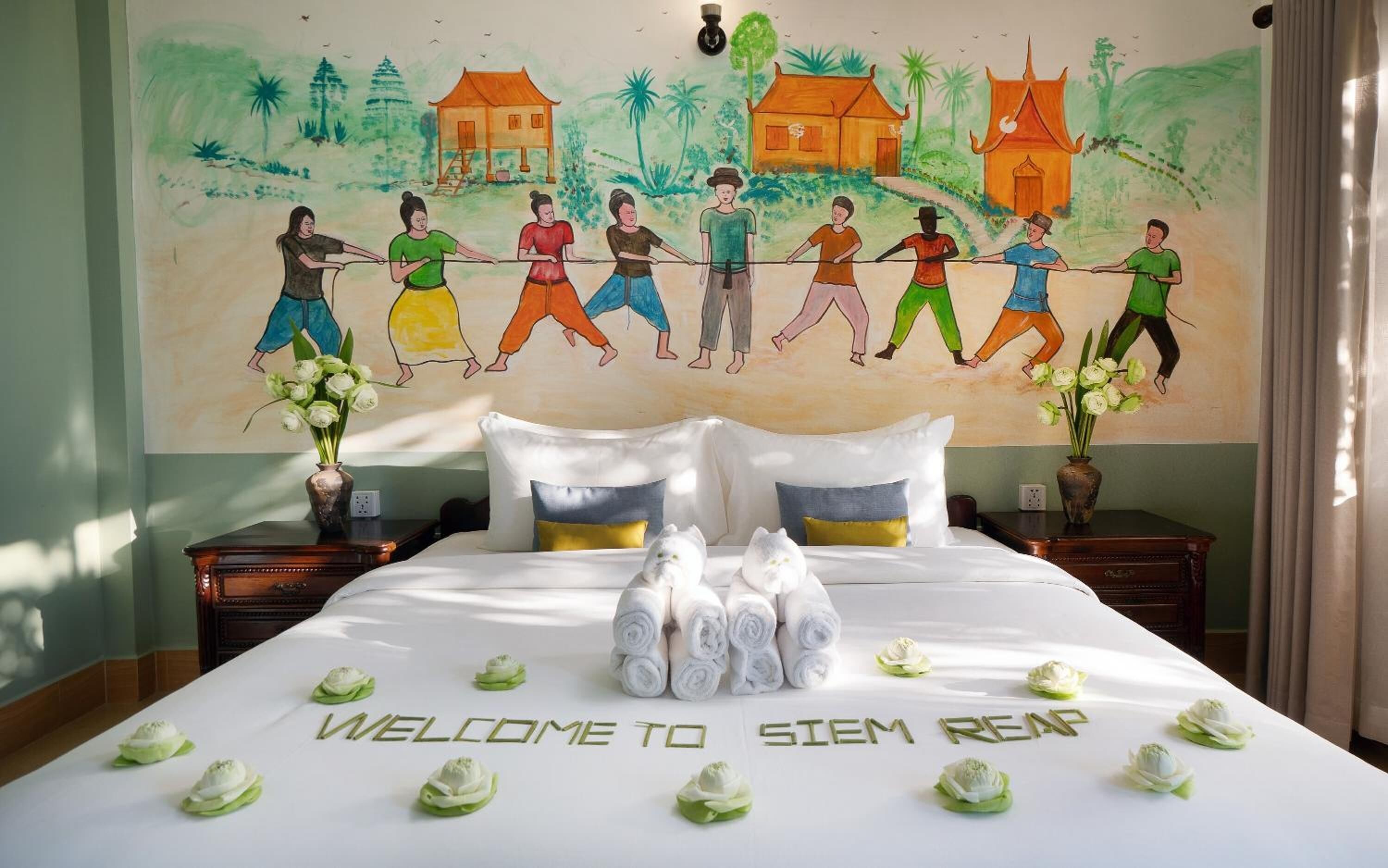 A hotel room with a bed, a mural, and a welcome sign on the bed.