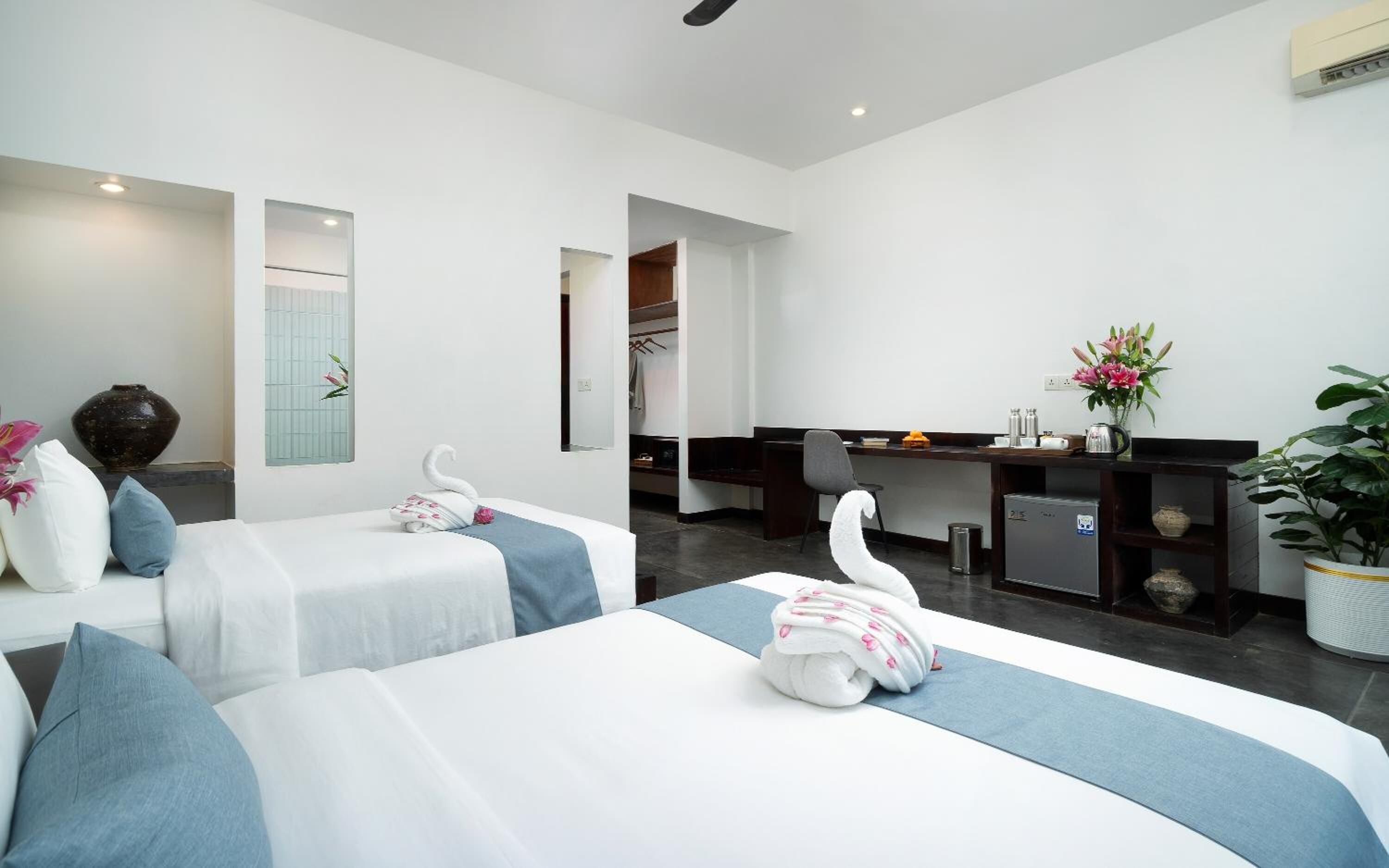A hotel room with two beds, a desk, a chair, a small fridge, and a potted plant.