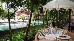 Une personne est assise sur un canapé en osier, près d’une table et d’une boisson, et a devant elle une piscine bordée de chaises longues et entourée de verdure.
