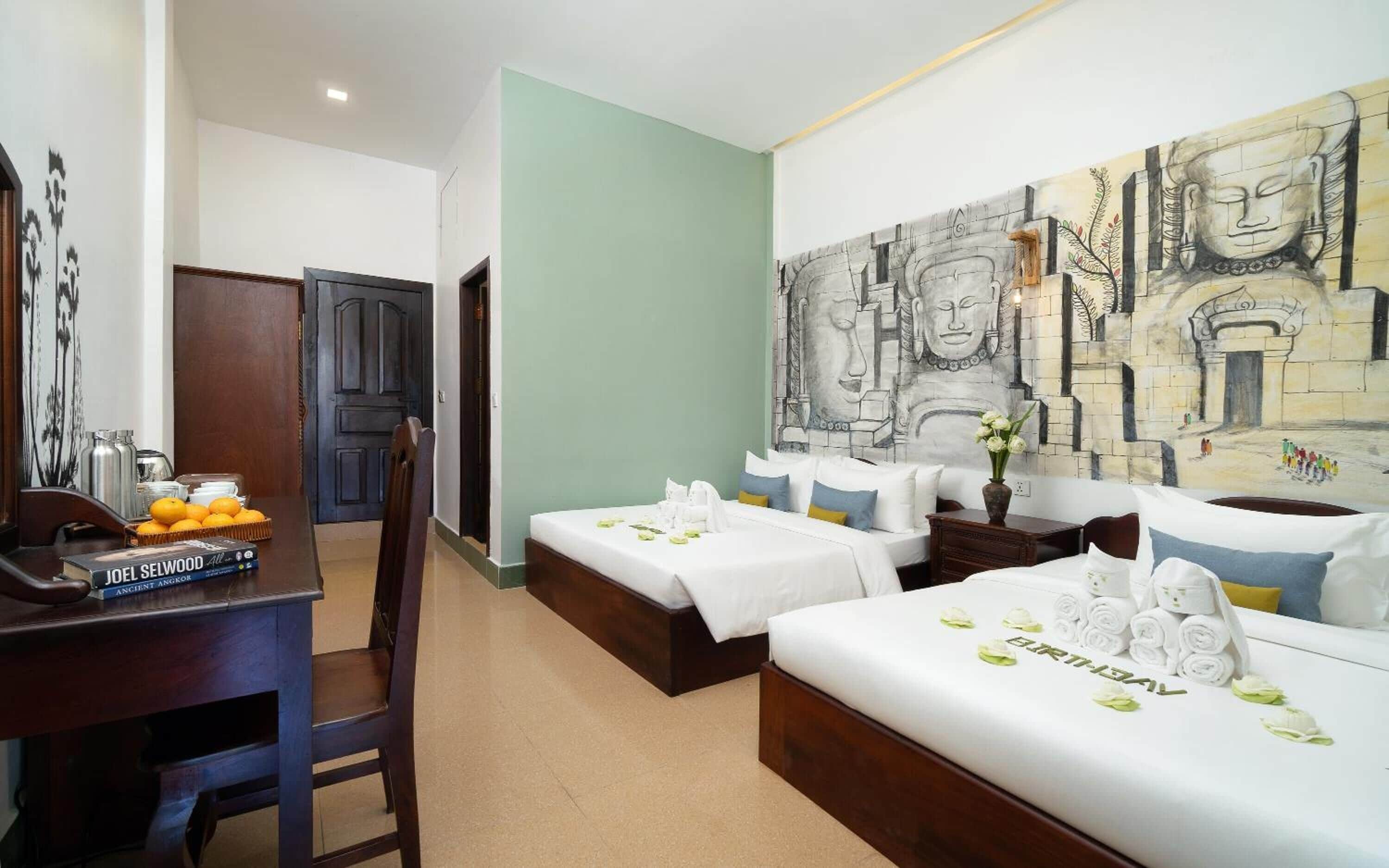A hotel room with a bed, a desk with a book, a chair, and a wall mural.