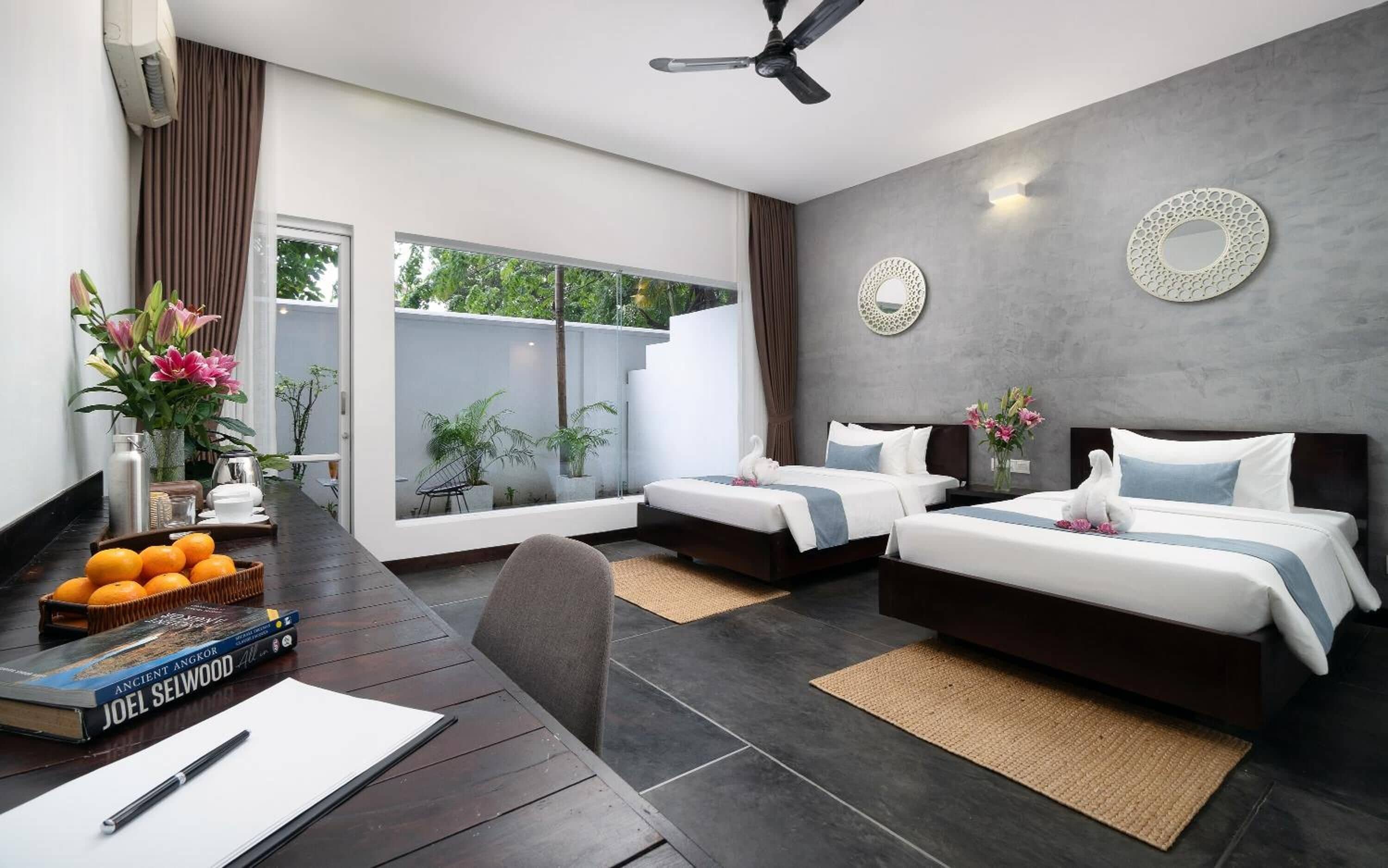 Una habitación de hotel moderna con dos camas, una zona de comedor con mesa y sillas, un ventilador de techo y un ventanal con vistas a zonas verdes.