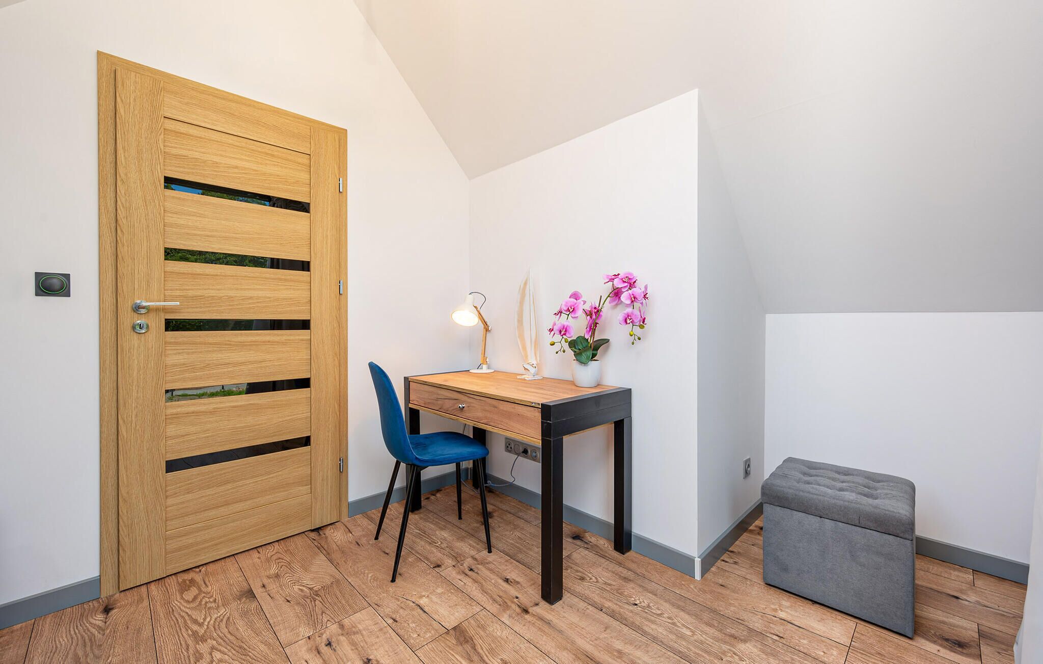 Een kamer met een houten deur, een bureau met stoel, een klein voetenbankje en een vaas met bloemen.