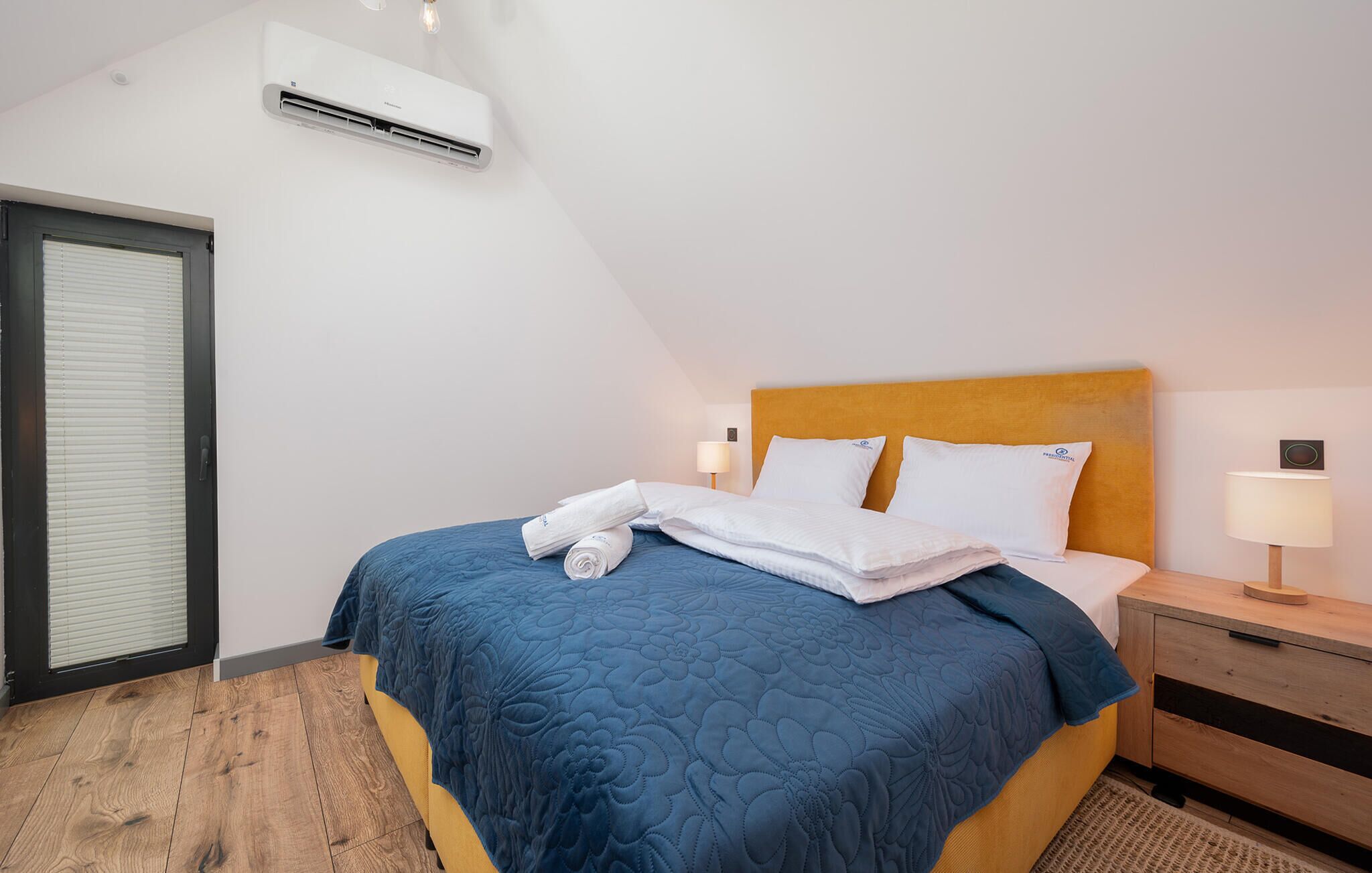 Ett hotellrum med en säng, ett sängbord, en lampa, en luftkonditioneringsanläggning och ett fönster med persienner.