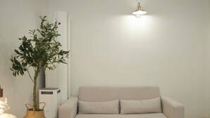A neutral-colored sofa with two cushions, a potted plant, a white lamp, and a white air purifier unit against a plain wall.