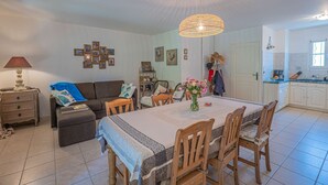 Un coin repas avec une table dressée pour quatre personnes, entourée de chaises en bois. Un lustre est suspendu au-dessus de la table, et on aperçoit un espace cuisine sur la droite.