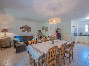 A dining area with a table set for four, surrounded by wooden chairs. There is a chandelier above the table, and a kitchen area is visible to the right. - VILLA SAINTE MAXIME - GARDEN - CLOSE TO THE CITY CENTER AND SHOPS (STE MAXIME)