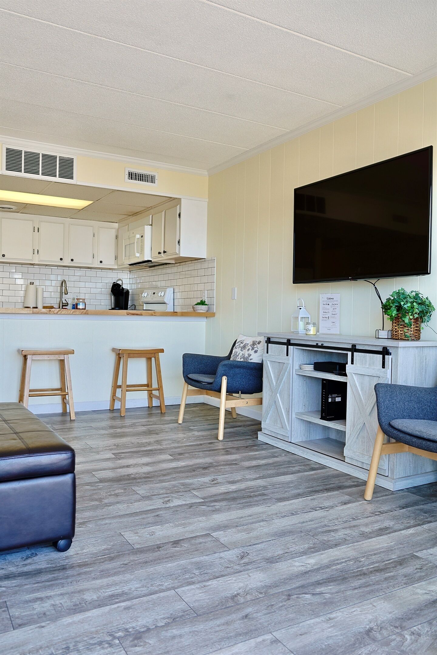 A modern kitchen with white cabinets, a large flat-screen TV, wooden stools, and a grey sofa.