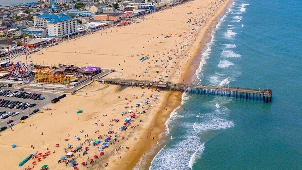 Vista aerea di una spiaggia con un molo che si estende nell'oceano, una ruota panoramica e una zona costiera affollata.