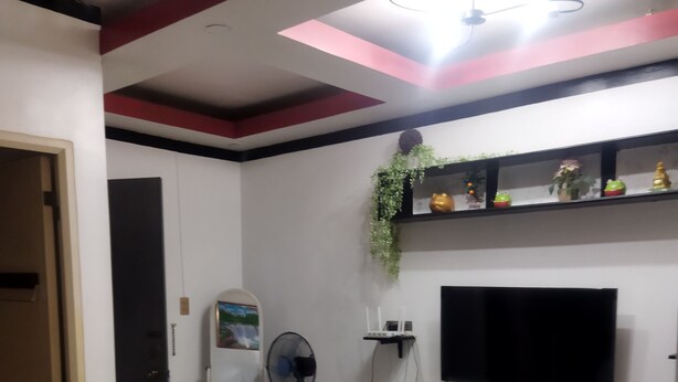 A living room with a TV mounted on the wall, a ceiling fan, a small table with a vase of flowers, and a sofa. - 1_Las Asturias Charming Condo Just Steps from Enchanted Kingdom (CITY OF SANTA ROSA)