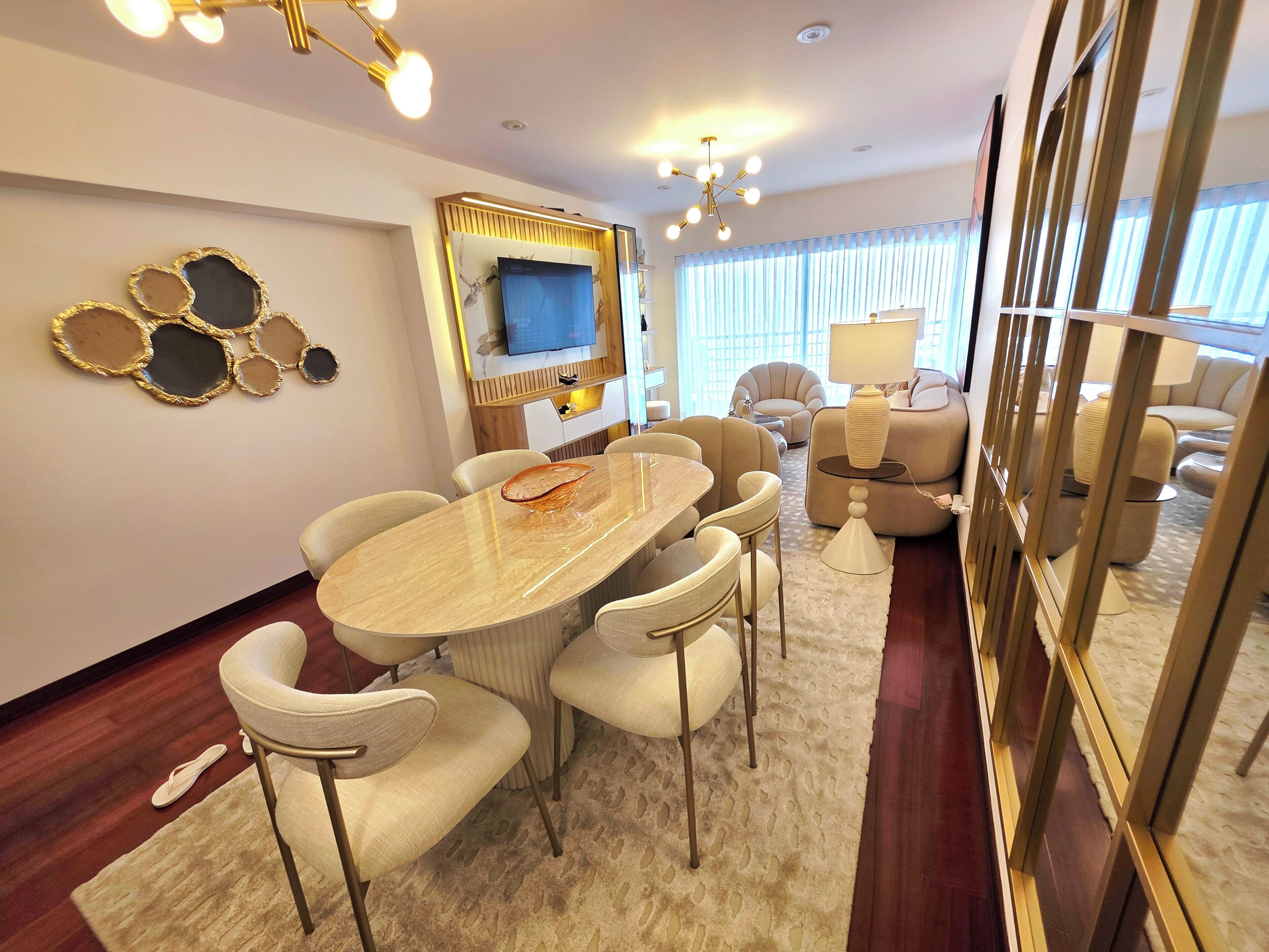 A modern living room with a marble coffee table, beige upholstered chairs, a television mounted on a wooden panel, and a large mirror reflecting the room.