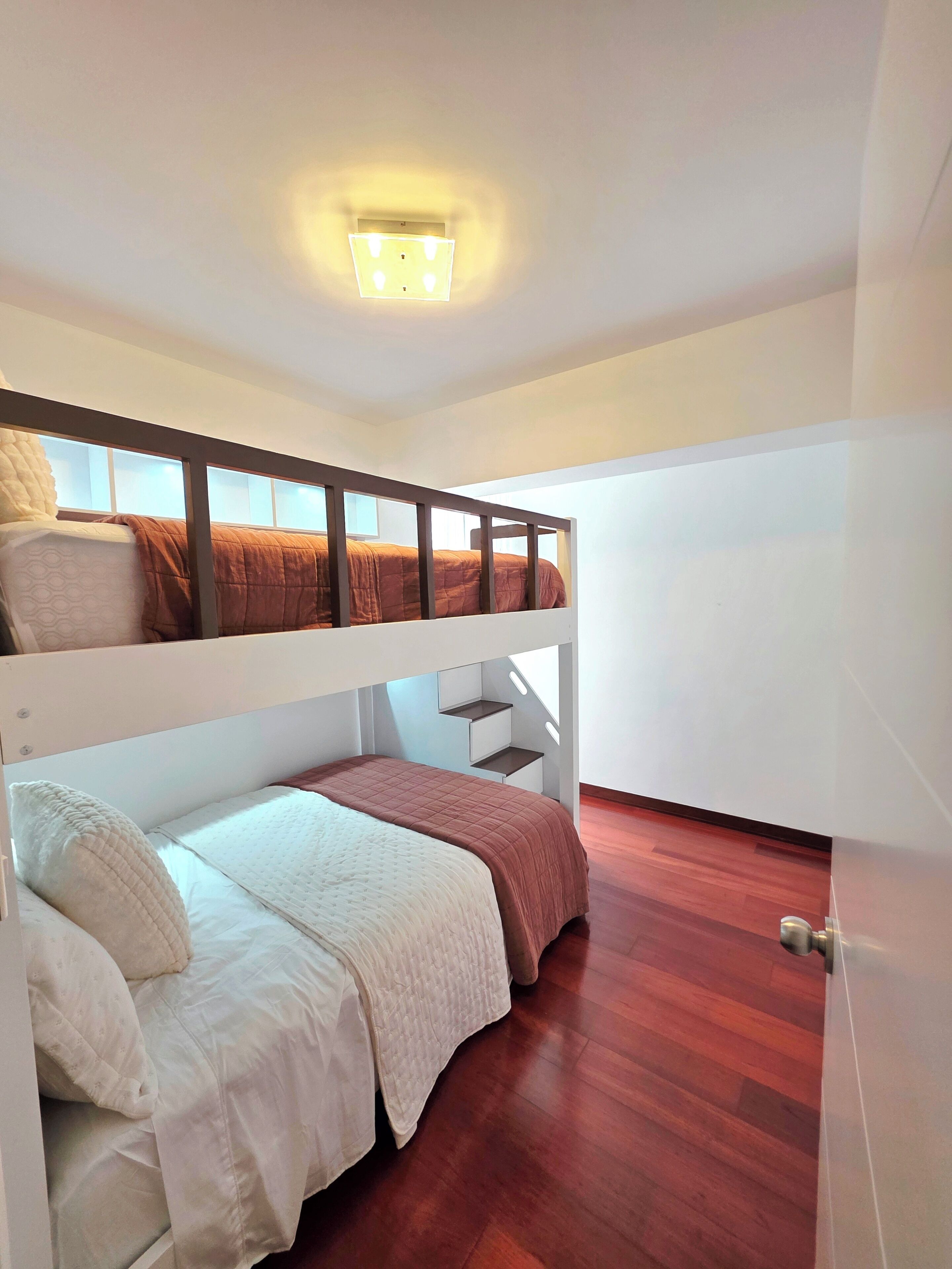A bunk bed room with a wooden floor, white walls, and a ceiling light fixture.