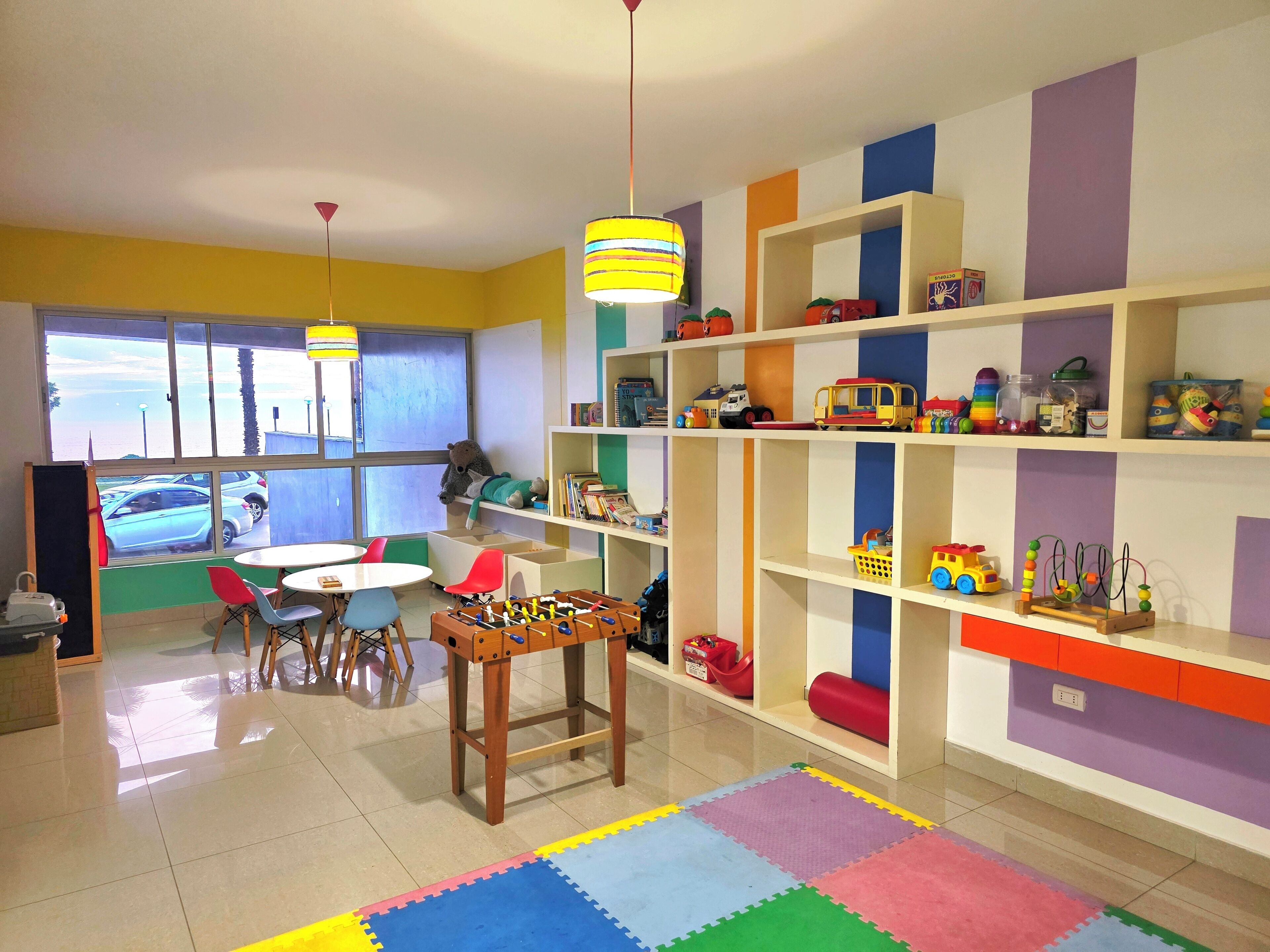 Une salle de jeux pour enfants avec un baby-foot, des étagères colorées et une fenêtre donnant sur l’extérieur.