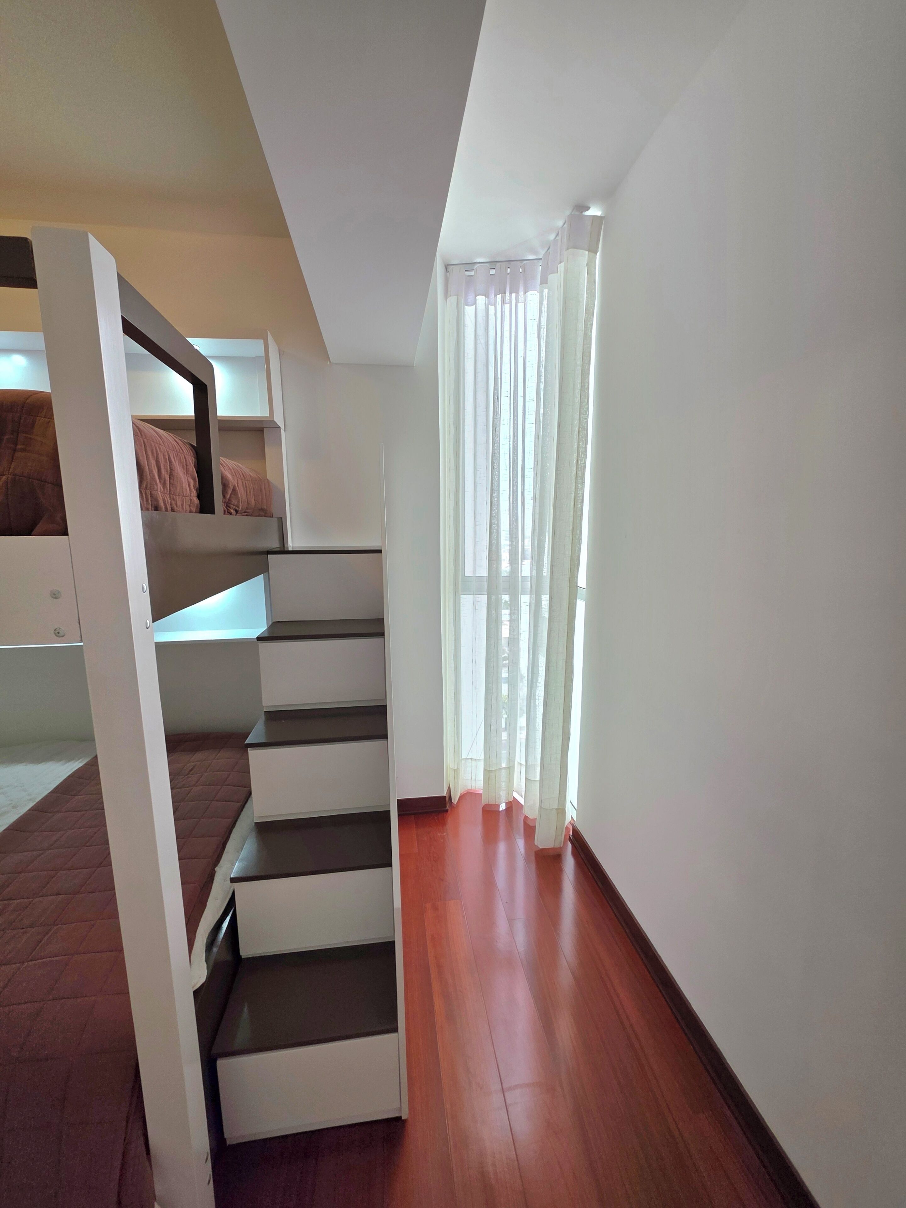 Quarto com beliche, escada e janela com cortinas leves.