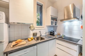Une cuisine moderne avec des meubles blancs, une hotte de cuisinière en acier inoxydable, un lave-vaisselle et une fenêtre avec des stores.
