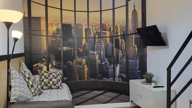 Ein Schlafzimmer mit Blick auf die Stadt, ein Bett mit gemusterten Betttextilien, eine Stehlampe, ein weißes Sideboard und eine Treppe.