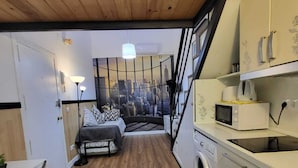 Ein kompakter Wohnraum mit Bett, Küchenzeile, Waschmaschine und Wandgemälde mit Stadtansicht.