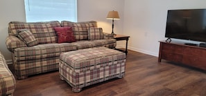 A living room with a plaid sofa set, a matching ottoman, a TV on a wooden stand, and a lamp on a side table.