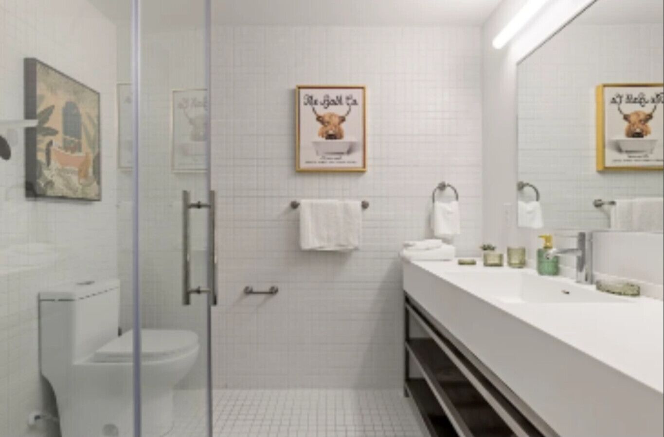 A modern bathroom with a glass shower enclosure, white tiled walls, and a long vanity with a mirror.
