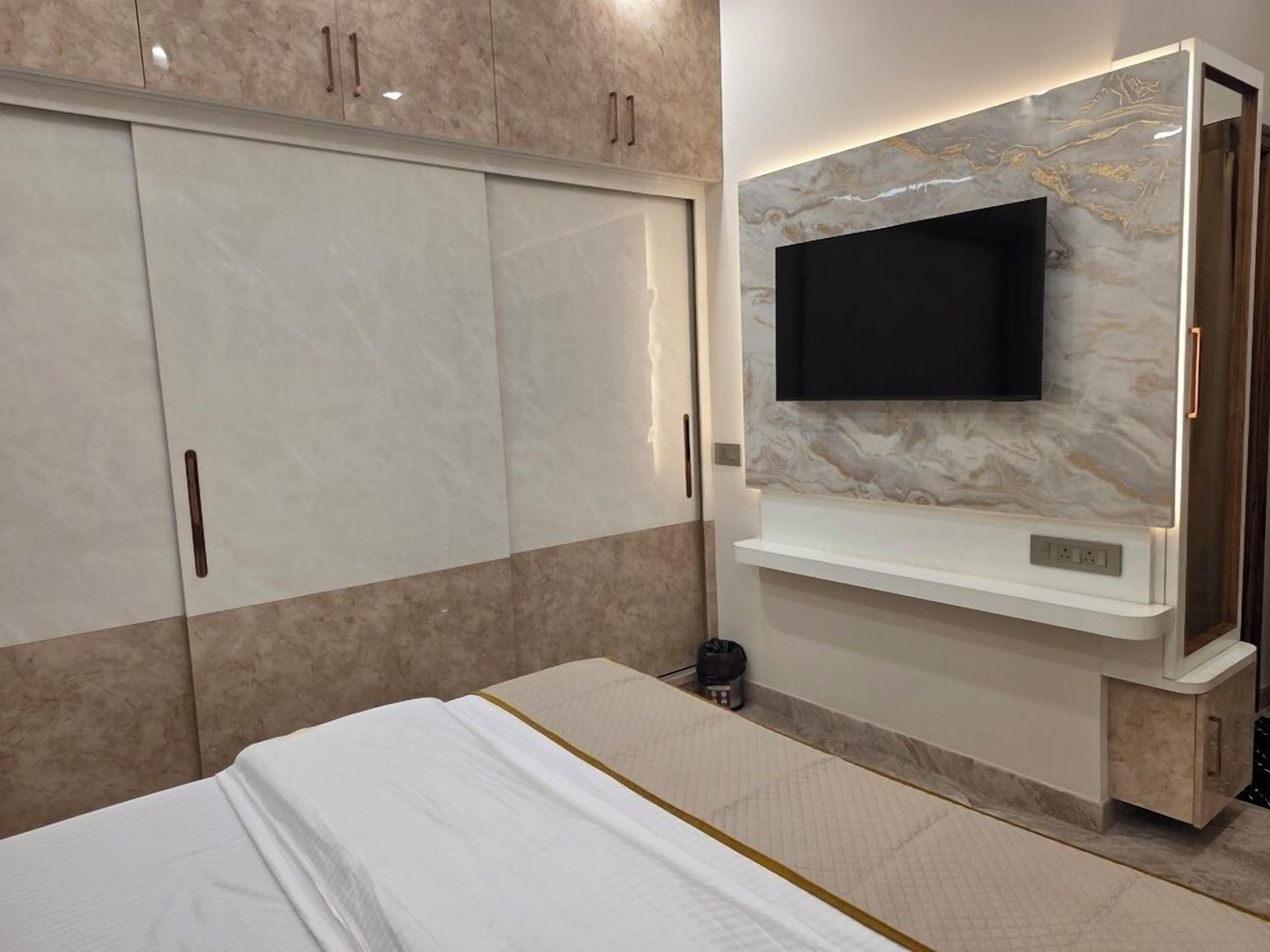 A bedroom with a bed, a wardrobe, a TV mounted on a marble wall, and a small shelf.