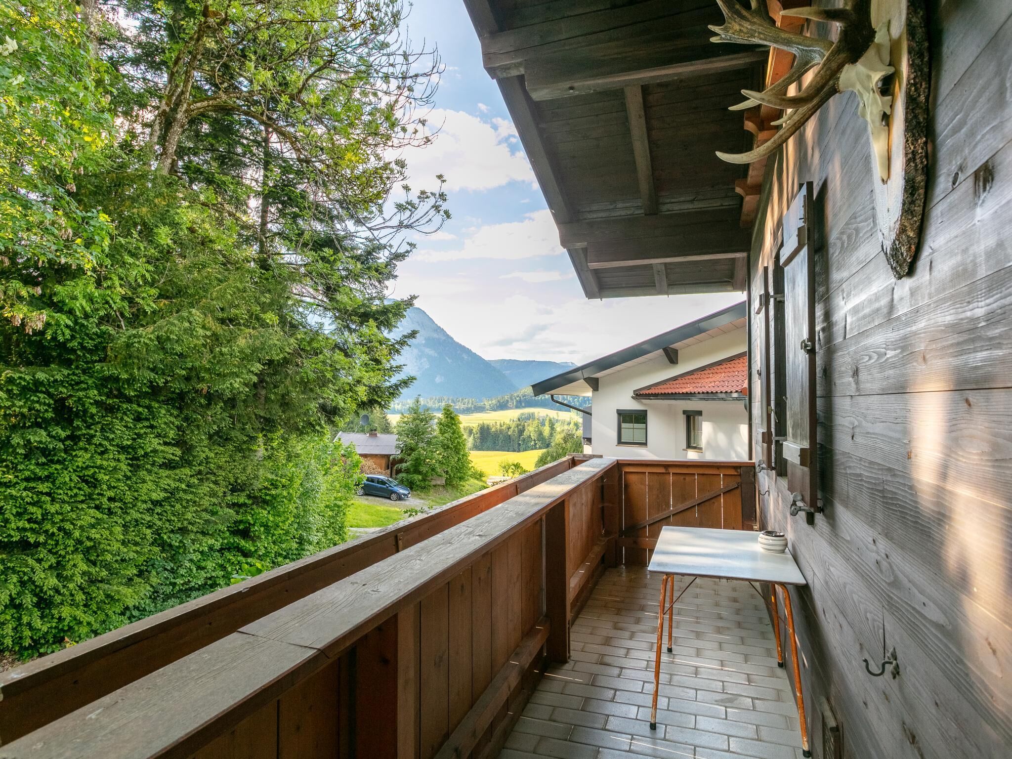 Ein Holzbalkon mit Tisch und Stühlen, der auf eine gebirgige Landschaft und ein Haus mit roten Ziegeldach blickt.