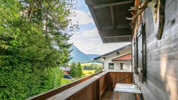 Ein Holzbalkon mit Tisch und Stühlen, der auf eine gebirgige Landschaft und ein Haus mit roten Ziegeldach blickt.
