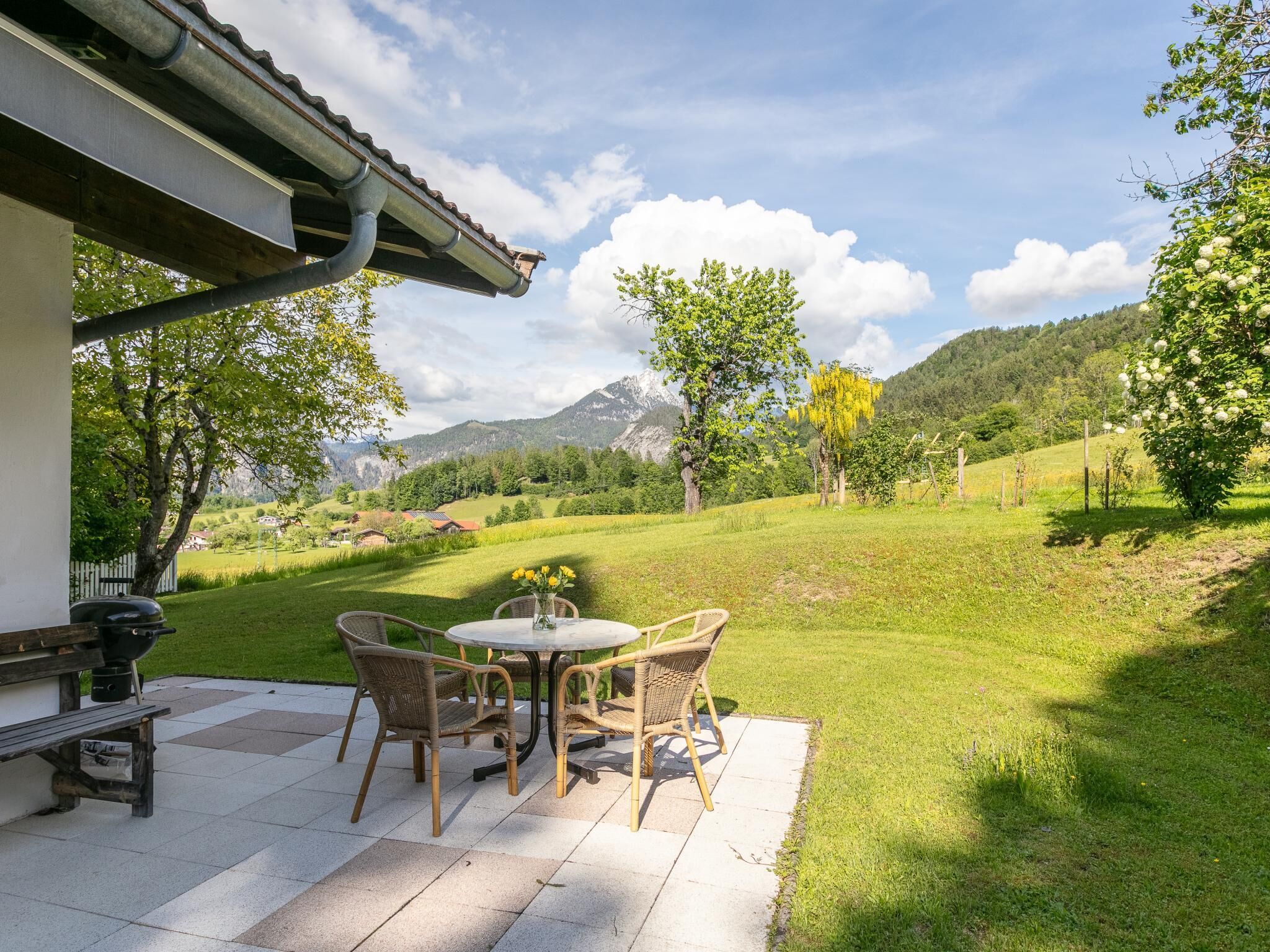 Eine Terrasse im Freien mit Tisch, Stühlen, einem Grill und Blick auf eine grüne Landschaft mit Bäumen und Bergen.