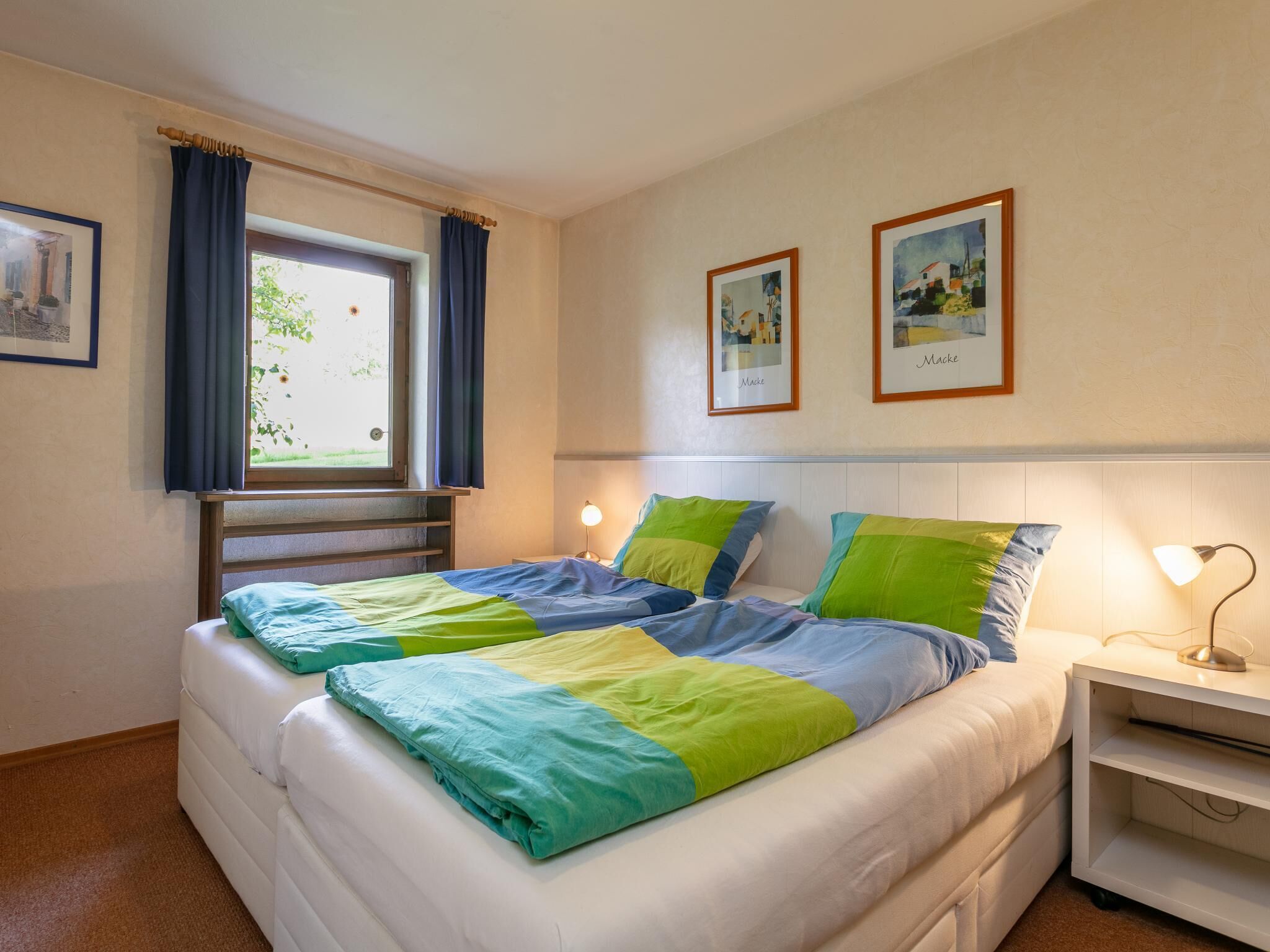 A double bed with a blue and green bedspread, a window with curtains, two framed pictures on the wall, and a bedside table with a lamp.
