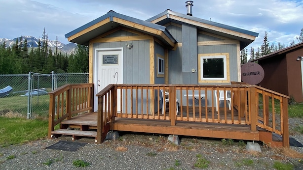 A small, single-story cabin with a wooden deck and a door, surrounded by a gravel driveway and a fenced area.