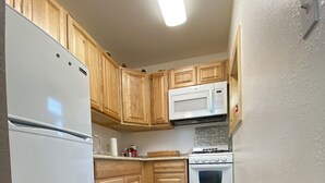 A compact kitchen with wooden cabinets, a white refrigerator, a microwave, an oven, and a small rug on the floor.