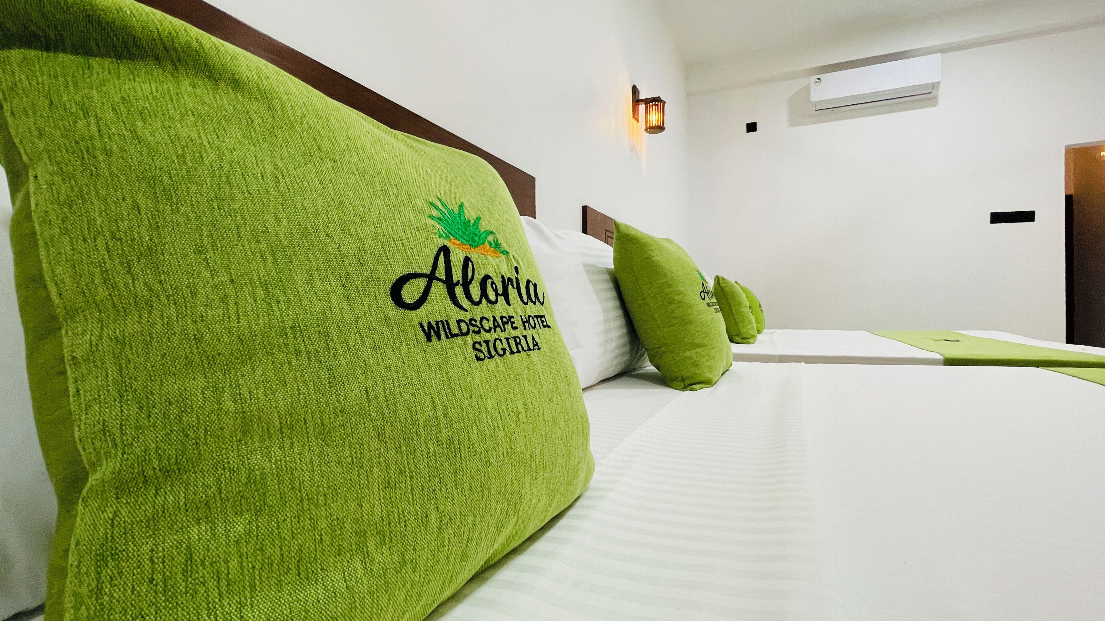 Quarto de hotel com cama, almofadas verdes, cabeceira, luminária fixada na parede e unidade de ar condicionado.