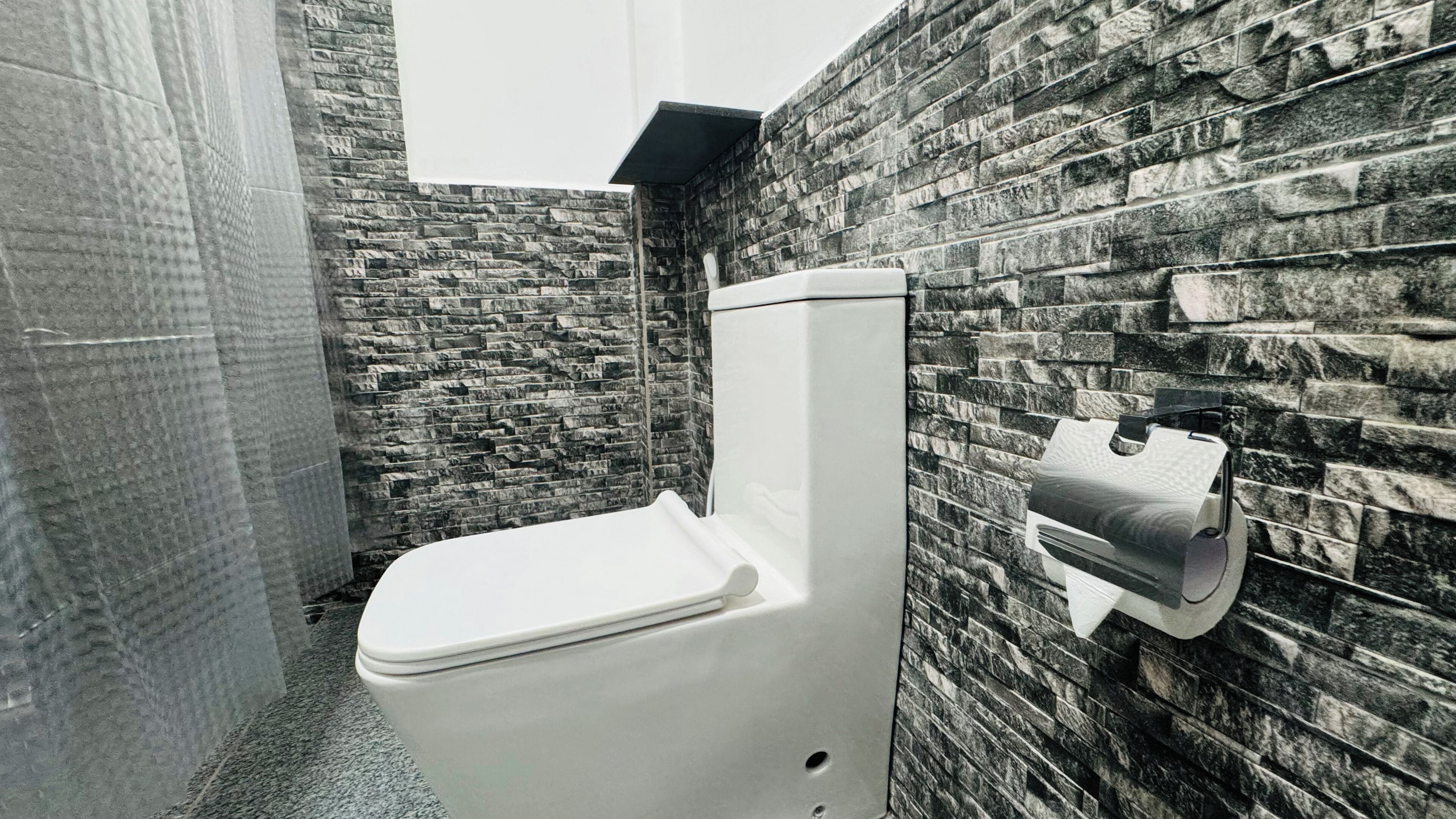 Un baño con inodoro blanco, pared con textura de piedra y dispensador de toallas de papel.