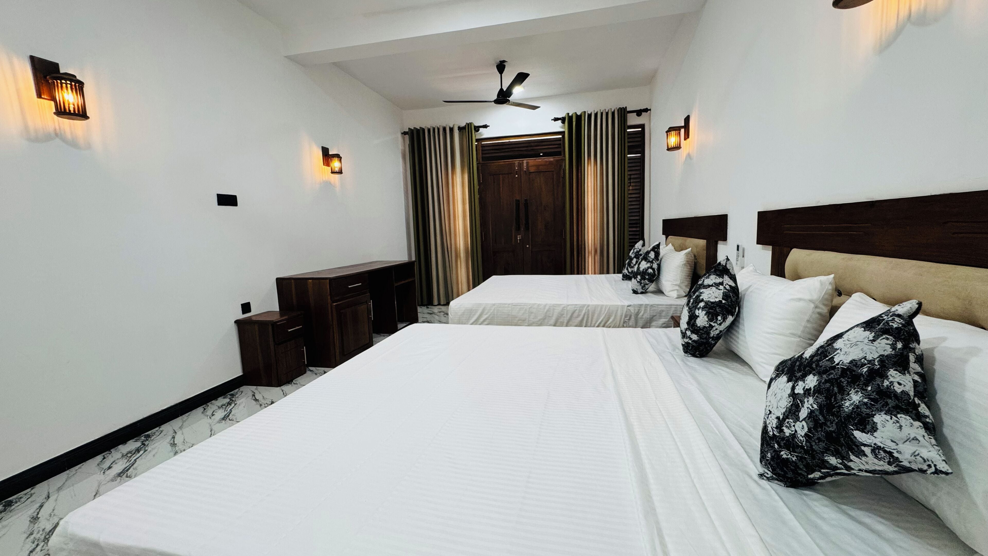 A hotel room with two beds, a ceiling fan, a wardrobe, and wall-mounted lights.