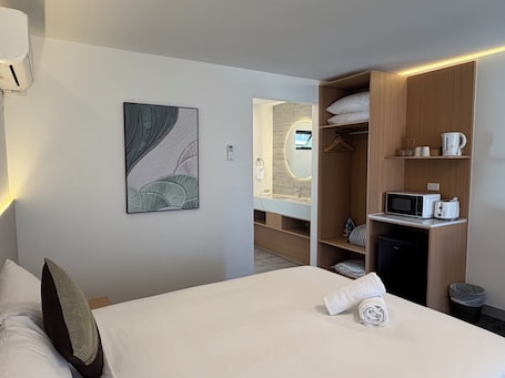 A hotel room with a bed, a bedside table with a rolled towel, a microwave, a toaster, a small shelf with cups and a kettle, a closet with hanging clothes, and a wall-mounted mirror.