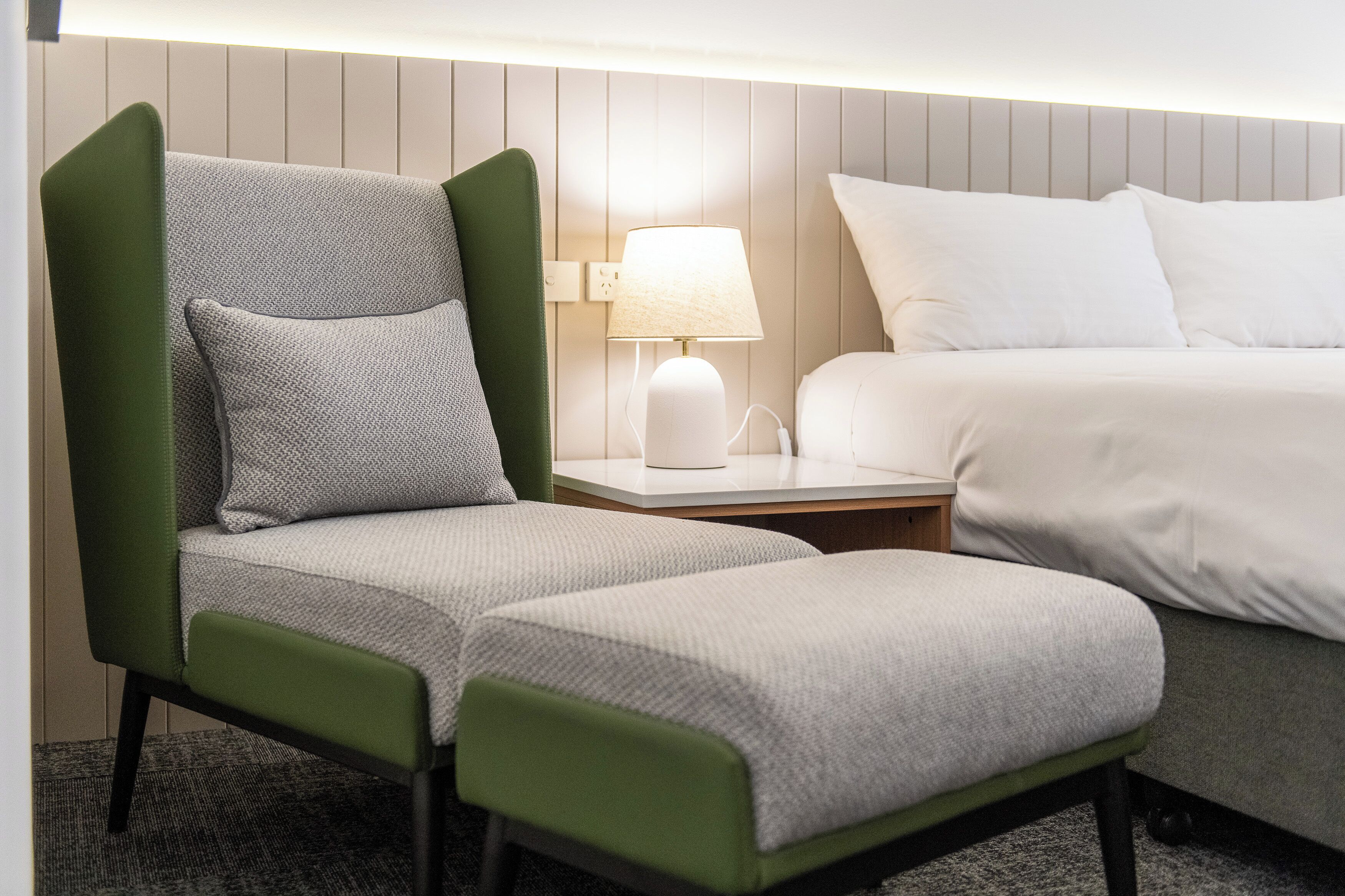 Une chambre d’hôtel avec un fauteuil à revêtement vert, une table de chevet blanche avec une lampe, et un lit avec une literie blanche.