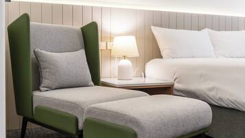 Une chambre d’hôtel avec un fauteuil à revêtement vert, une table de chevet blanche avec une lampe, et un lit avec une literie blanche.