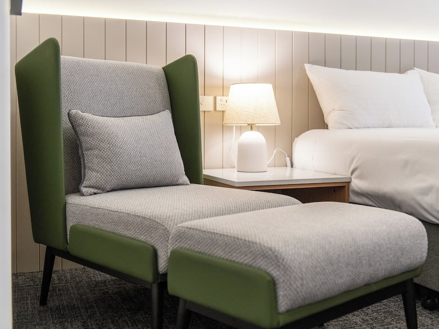 Una habitación de hotel con una butaca tapizada de verde, una mesita de noche blanca con una lámpara y una cama con ropa de cama blanca.