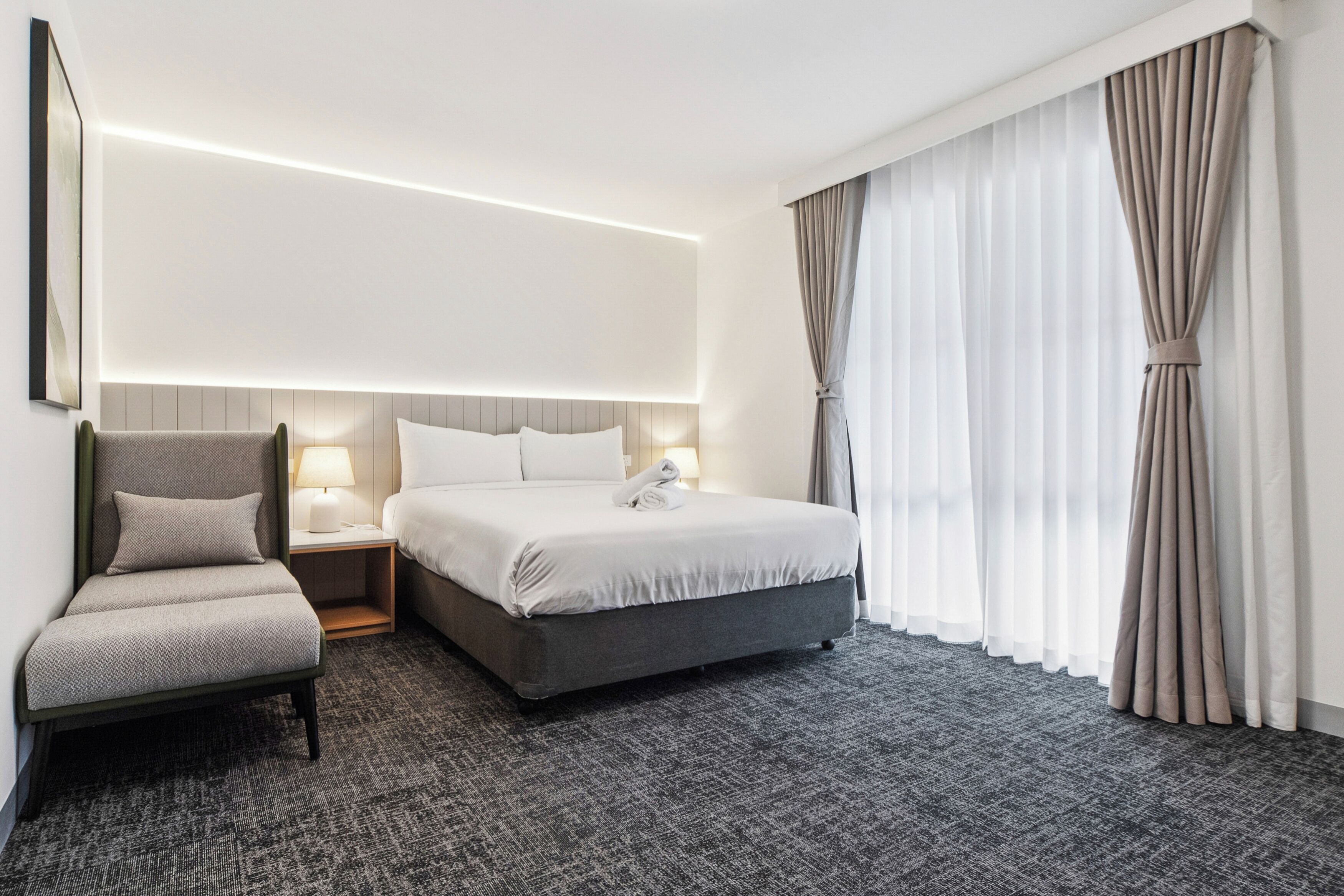 A hotel room with a bed, a chair, a nightstand, and a large window with curtains.