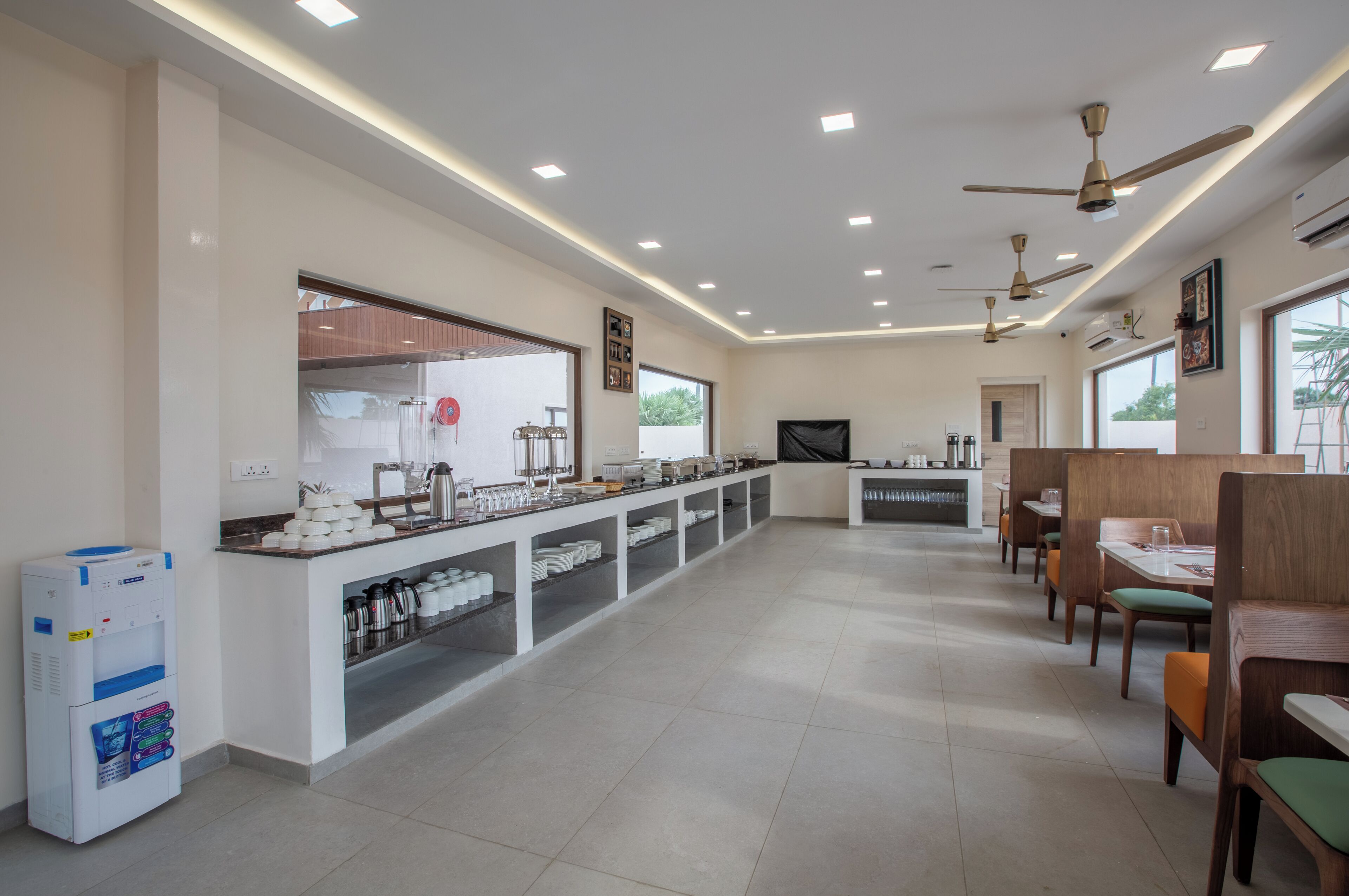 A spacious hotel dining area with a long counter, multiple ceiling fans, and a water dispenser.