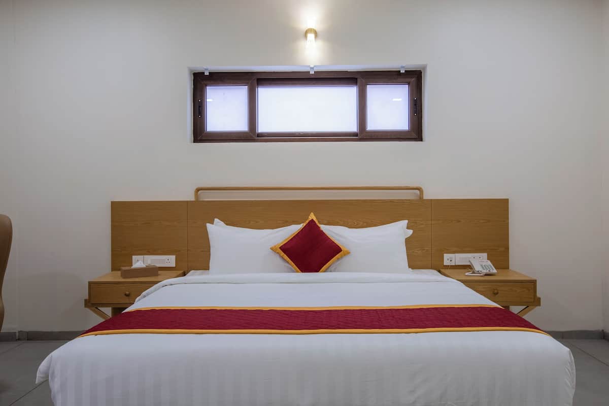 A hotel room with a wooden headboard, a bed with white bedding and a red accent pillow, a nightstand with a phone, and a window above the bed.
