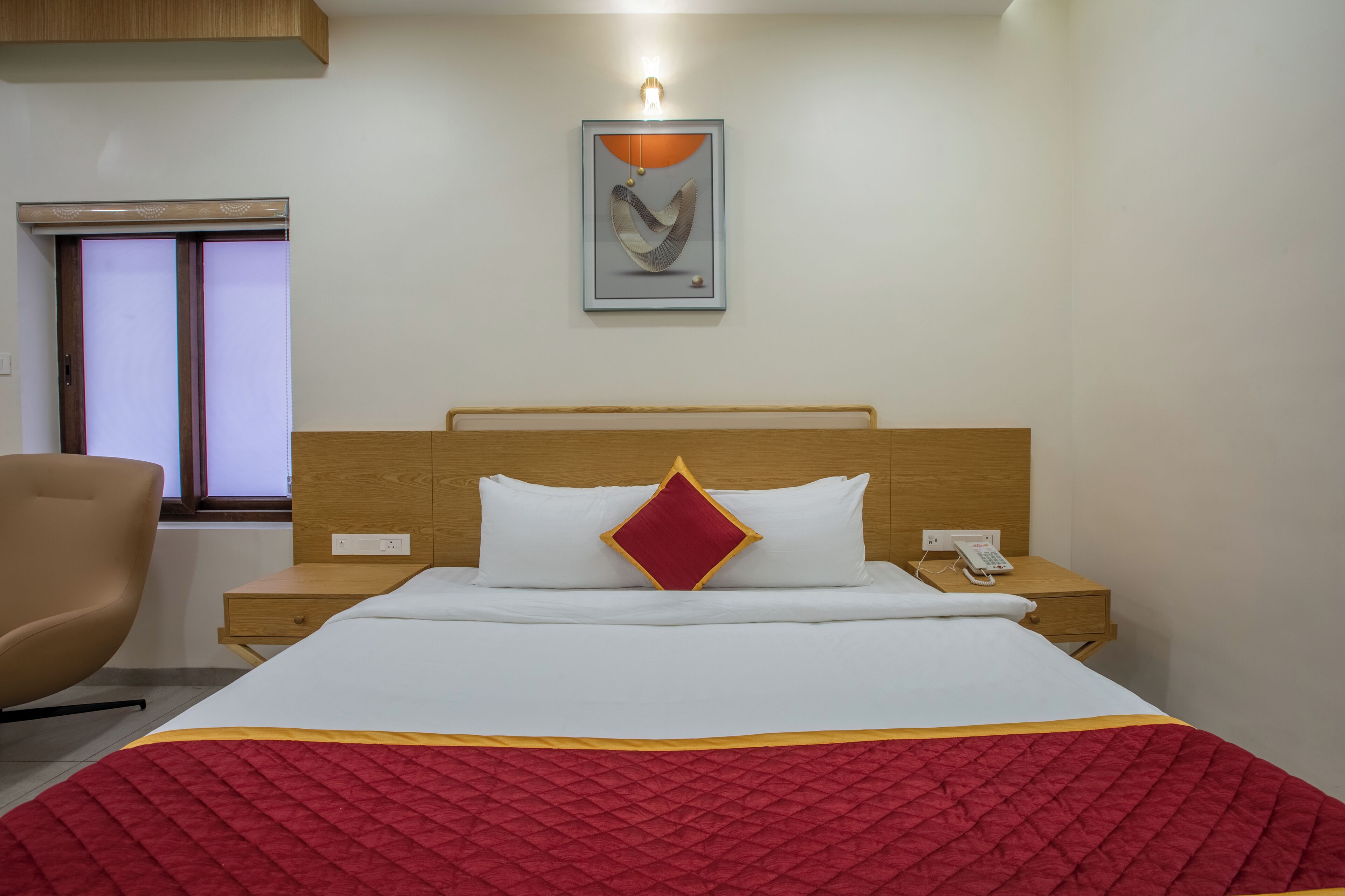 Una habitación de hotel con cama, mesita de noche, una silla, ventana y un cuadro colgado en la pared.