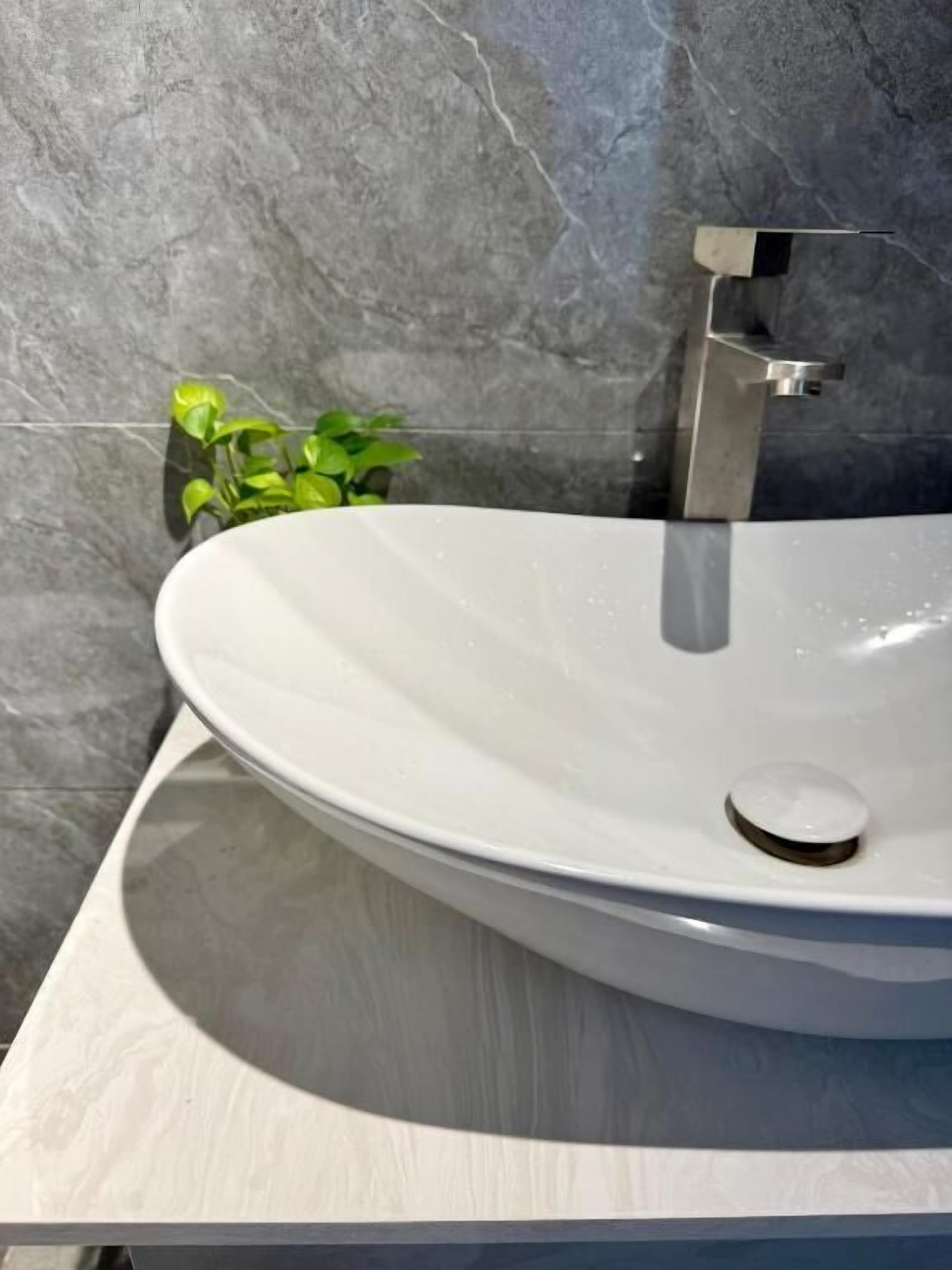 A white oval sink with a modern faucet, set against a grey tiled wall with a small green plant beside it.