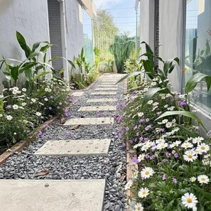 Un sentier bordé de pierres plates et agrémenté de différentes plantes, qui mène à un bâtiment doté de grandes fenêtres.