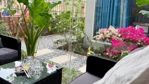Een kamer met een glazen tafel, een stoel, een potplant en uitzicht op een tuin met roze bloemen en rode, bolvormige decoraties.