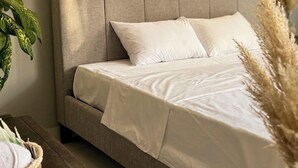 Een bed met een grijs gestoffeerd hoofdeinde, witte beddengoed en een macramé wanddecoratie.