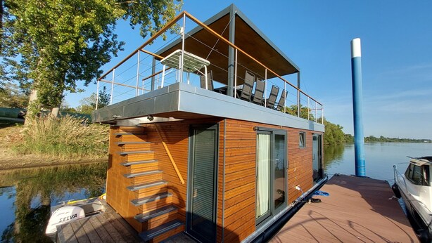 Une maison flottante moderne, avec une façade en bois, un balcon doté d’un espace salon et un bateau amarré à proximité.
