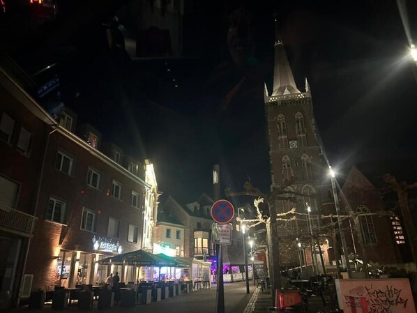 Eine nächtliche Straßenszene mit einem markanten Kirchturm, Außenbereich mit Sitzgelegenheiten und beleuchteten Gebäuden.