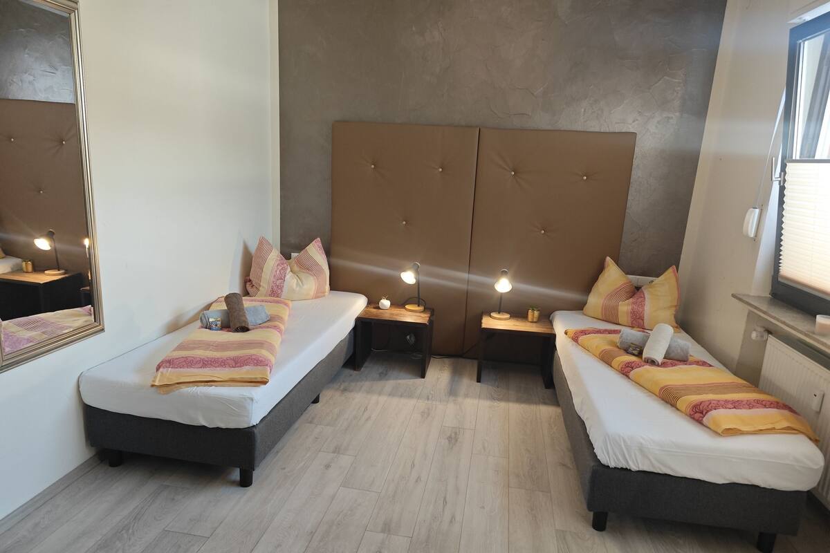 A hotel room with two beds, a large padded headboard, bedside tables with lamps, and a mirror.